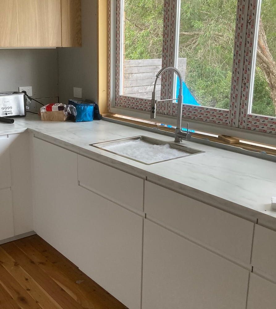 Modern White Kitchen With a Sink by a Window and Light Wood Cabinets — Corrimal Plumbing Services in Austinmer, NSW