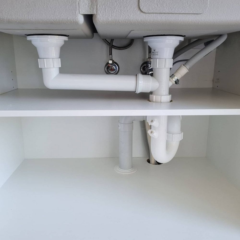 White kitchen sink plumbing under cabinet with pipes and drain. — Corrimal Plumbing Services in Bulli, NSW