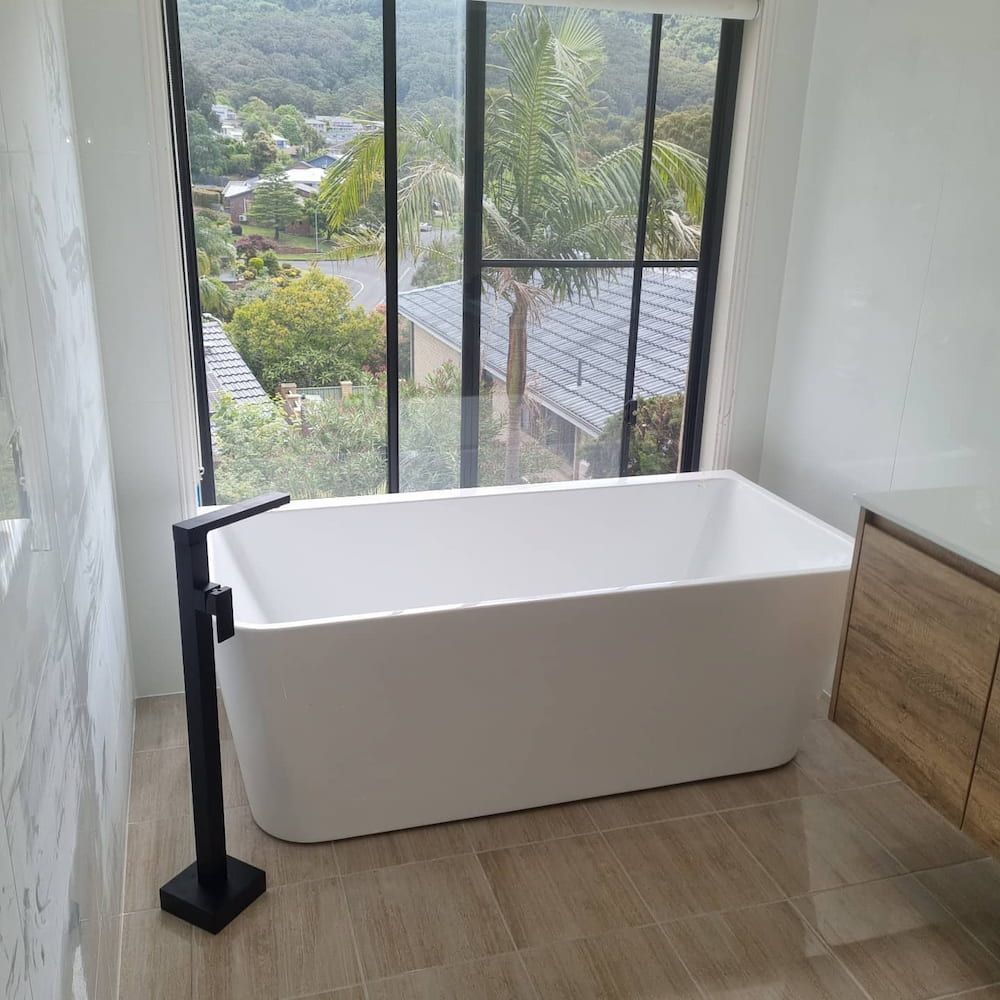 Modern White Bathtub Near a Large Window, Black Faucet and Wooden Vanity — Corrimal Plumbing Services in Thirroul, NSW