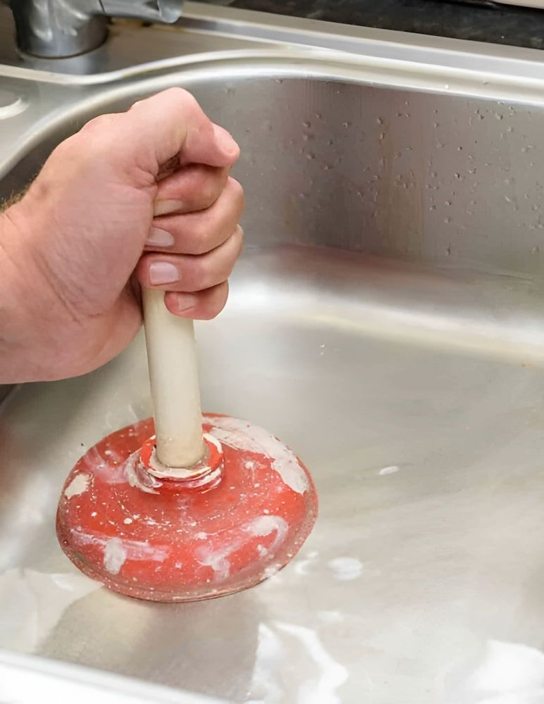 Hand Holding a Red Plunger in a Stainless Steel Sink — Corrimal Plumbing Services in Shellharbour, NSW