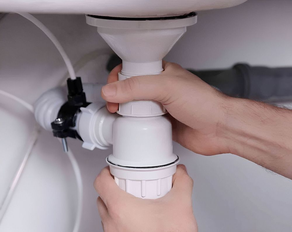 Hands Assembling a White Plastic Sink Drain Under a White Sink — Corrimal Plumbing Services in Austinmer, NSW