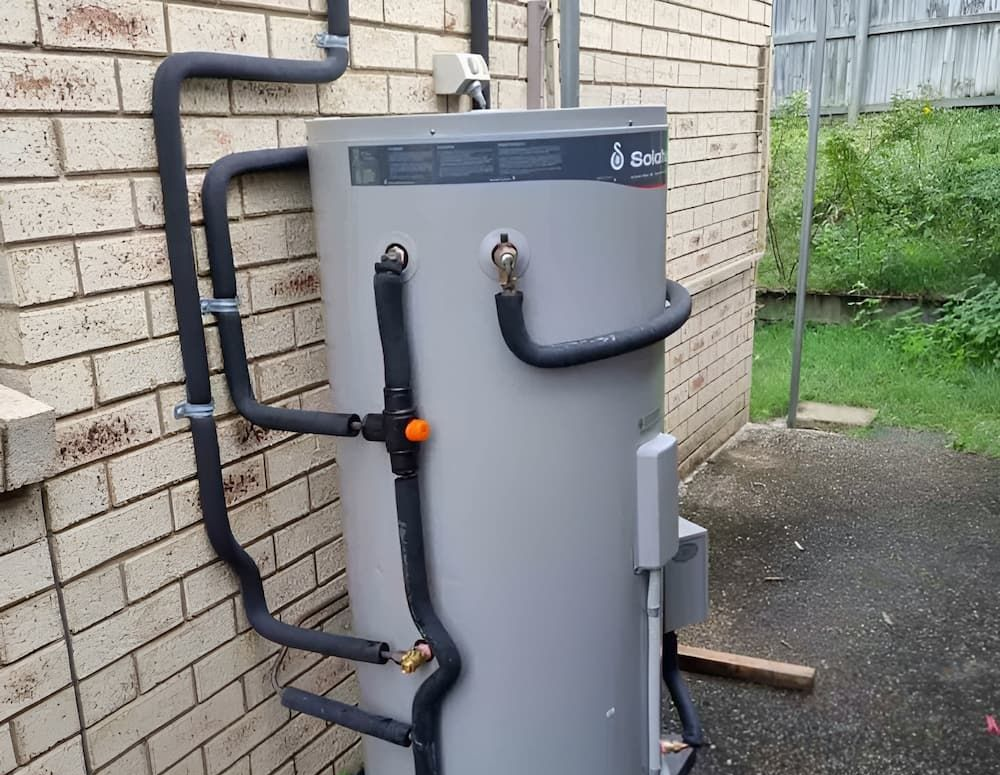 Grey Water Heater Against a Brick Wall, With Black Insulated Pipes — Corrimal Plumbing Services in East Corrimal, NSW